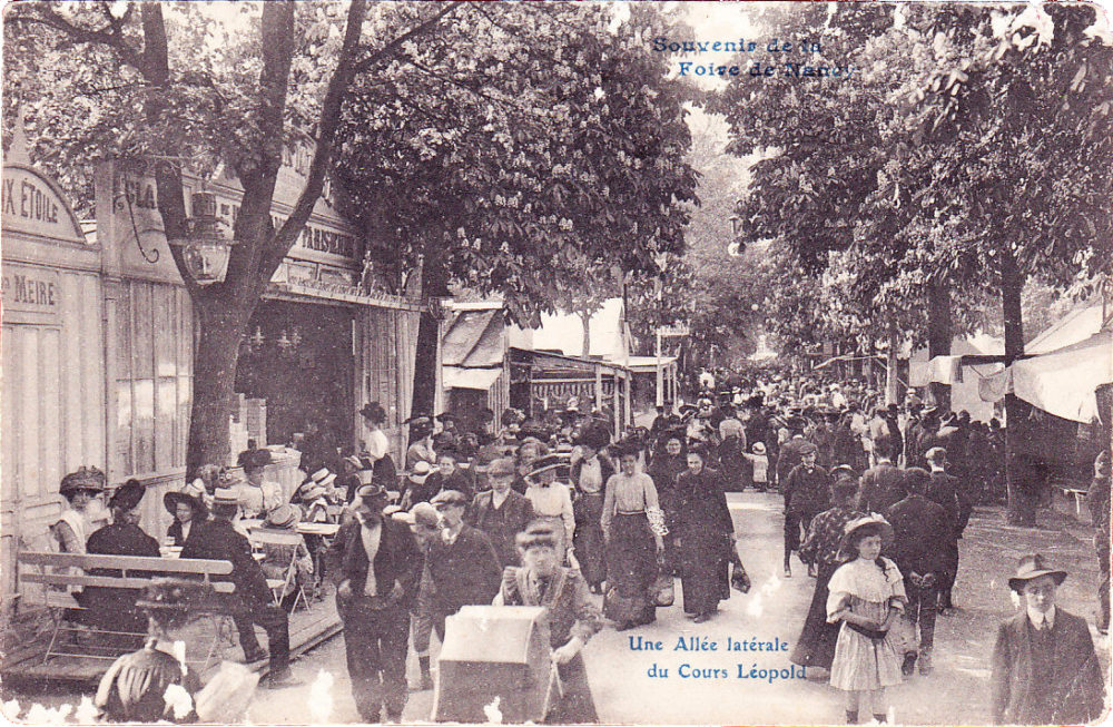 Carte postale ancienne où l'on voit la foule se presser entre les baraques, dans une allée bordée d'arbres